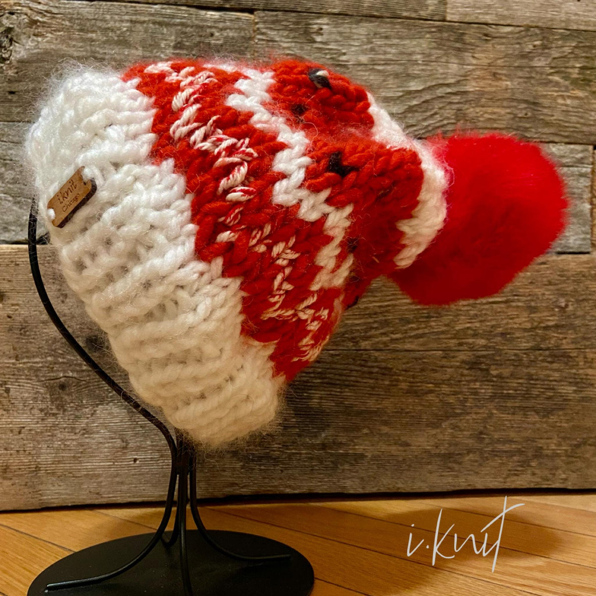May include: A white and red knit beanie with a red pom pom. The beanie has a white knit band and a red knit pattern. The beanie is on a black stand. The beanie has a small brown tag that says 'i.knit'.